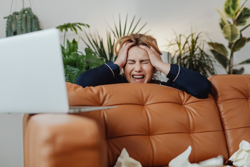 Woman upset and frustrated, hiding from her laptop behind a couch.